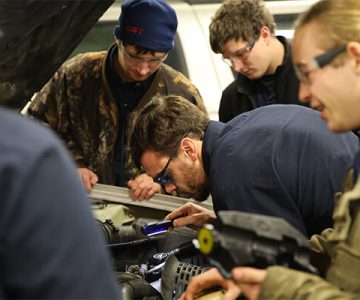 students holding a flashlight looking into engine of a car
