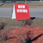 Image of red now hiring sign sitting by road outside business