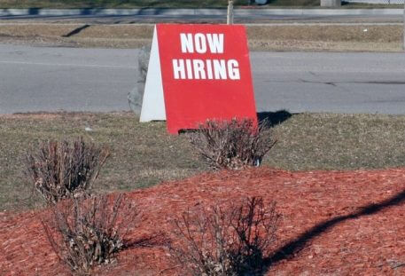 Image of red now hiring sign sitting by road outside business