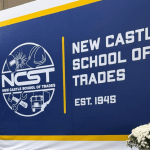 Navy blue backdrop reading New Castle School of Trades with vases of flowers