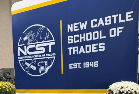 Navy blue backdrop reading New Castle School of Trades with vases of flowers