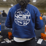 NCST student standing at career fair table, NCST emblem on back of his jacket