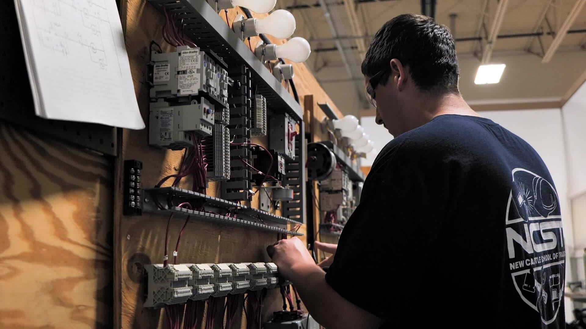Josh wearing safety glasses works on an electrical training board, connecting wires and components in a hands-on lab or classroom environment.