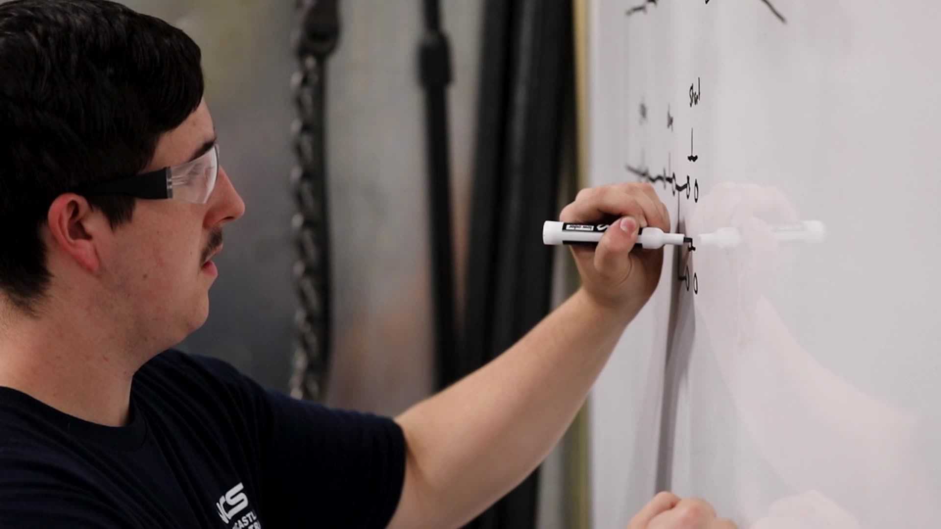 Josh wearing safety glasses writes notes or a diagram on a whiteboard with a marker in a workshop or classroom setting.