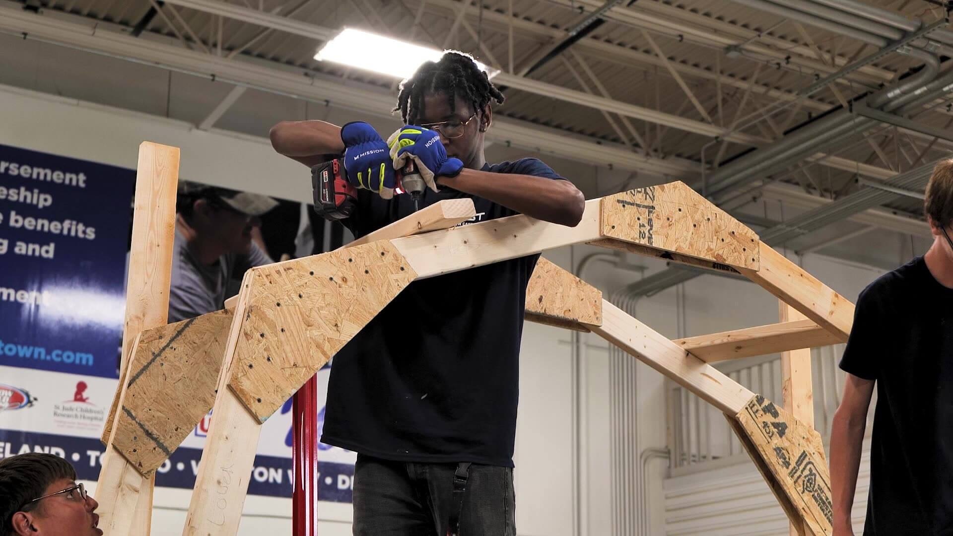 Benjamin Soto wearing work gloves uses a power drill to assemble a wooden roof truss structure