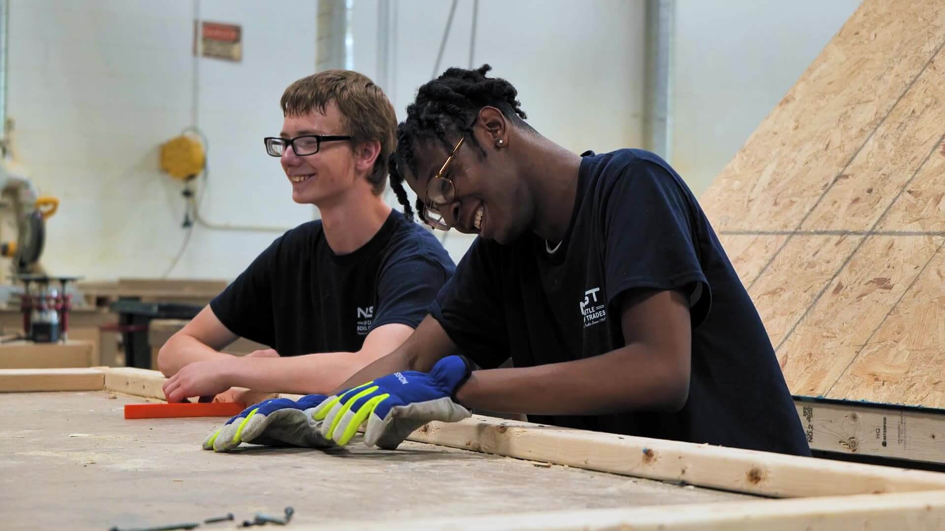 Two smiling trade school students wearing work gloves work together on a wood framing project
