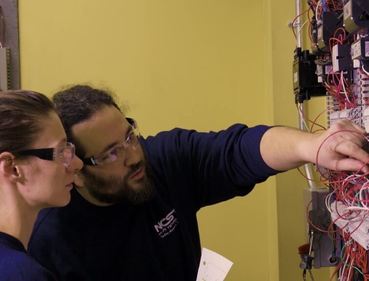 Electrical instructor wearing safety glasses points out wiring connections on a control panel to a student