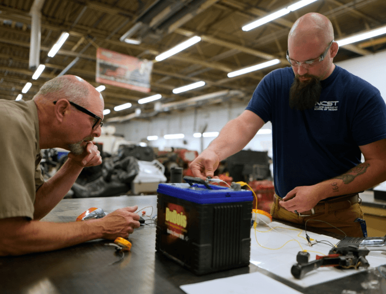 An NCST instructor and a student working through an electrical exercise with a battery and wiring on a workbench in a powersports repair shop.
