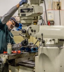 Machining student in a green uniform, cap, and safety glasses adjusting the head of a vertical milling machine