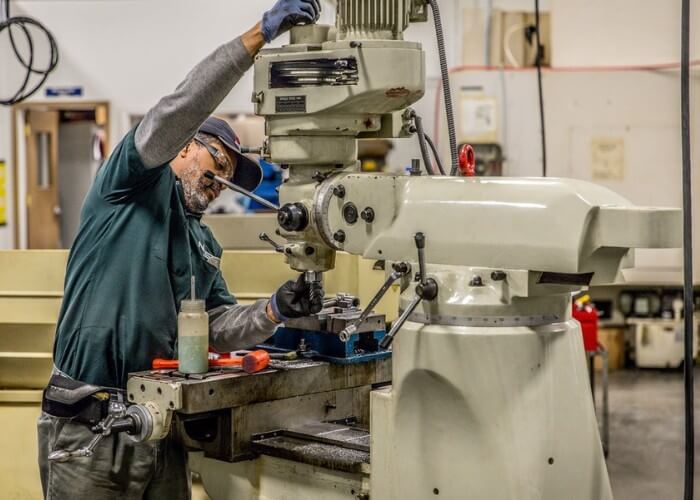 Machining student in a green uniform, cap, and safety glasses adjusting the head of a vertical milling machine