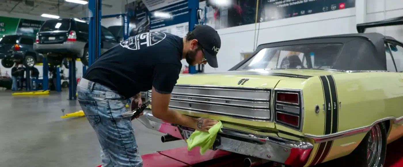 NCST student polishing the rear bumper of a classic yellow Dodge convertible in a shop, with vehicles on lifts in the background.