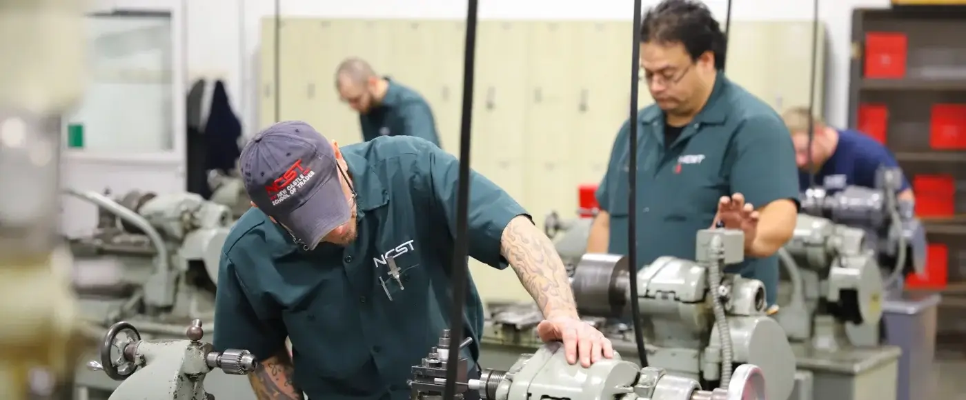 NCST students operating metal lathes in a machining shop, with lockers and tool cabinets in the background.