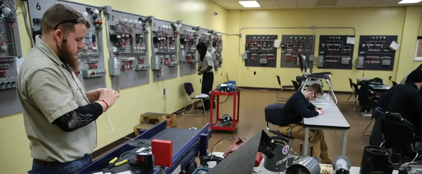 Students practicing wiring at electrical trade school stations, with numbered practice panels of switches, breakers, and lightbulbs lining the walls.