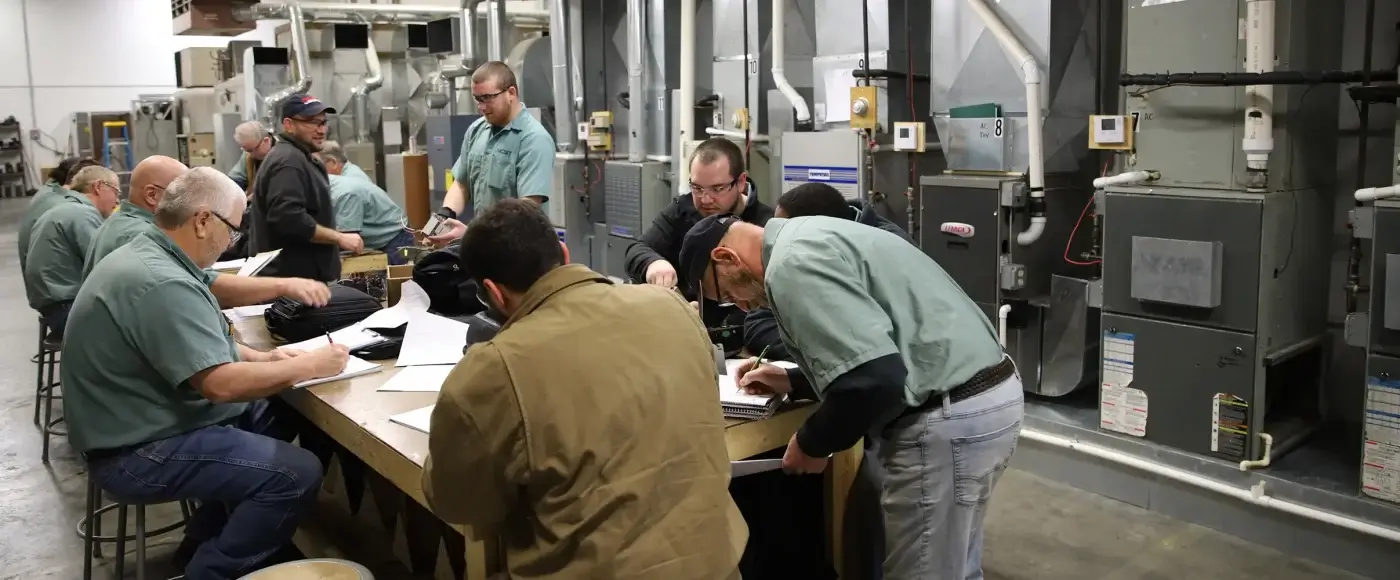 NCST students in uniforms gathered around a workbench taking notes and examining components, with furnaces, ductwork, and heating units filling the lab.