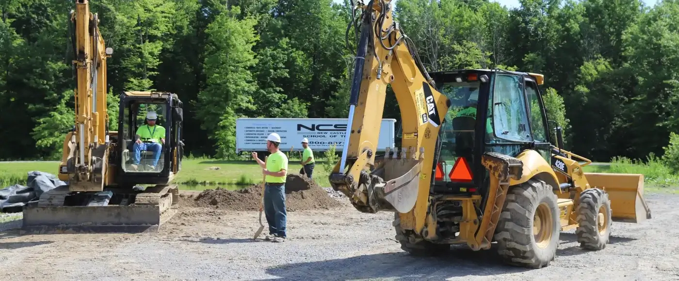 Heavy equipment operator students in safety gear training with a CAT excavator and backhoe at an outdoor NCST site, with a school trailer and trees in the background.