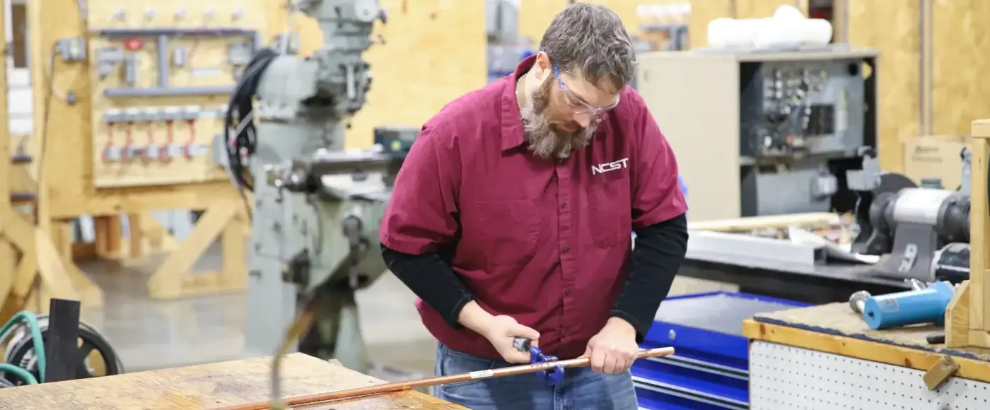 NCST student wearing safety glasses using a tube bender on a length of copper pipe, with a milling machine and electrical panels in the shop behind him.