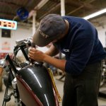 A motorcycle repair student in an NCST cap and sweatshirt working on the handlebars of a motorcycle in a shop