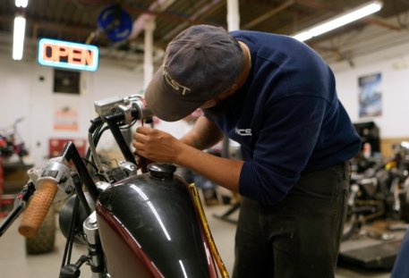 A motorcycle repair student in an NCST cap and sweatshirt working on the handlebars of a motorcycle in a shop