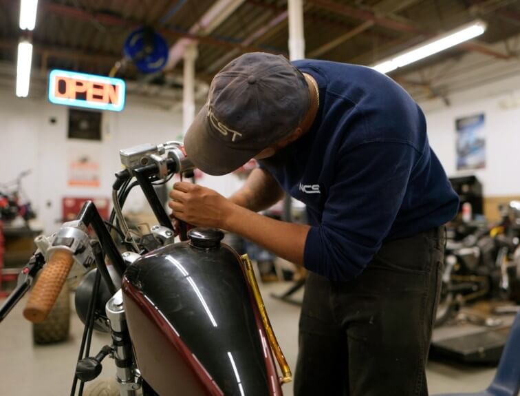 A motorcycle repair student in an NCST cap and sweatshirt working on the handlebars of a motorcycle in a shop