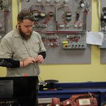 An electrical technician student in an NCST shirt and safety glasses working with wire at a bench, with wired practice control panels on the wall behind him.