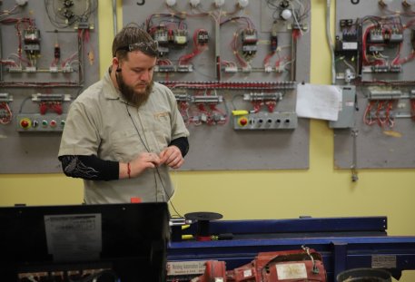 An electrical technician student in an NCST shirt and safety glasses working with wire at a bench, with wired practice control panels on the wall behind him.