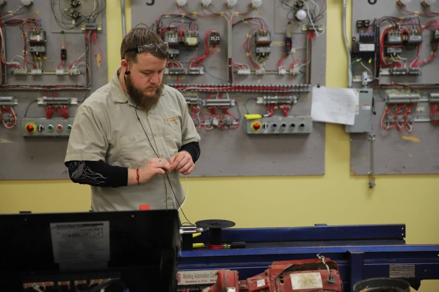 An electrical technician student in an NCST shirt and safety glasses working with wire at a bench, with wired practice control panels on the wall behind him.