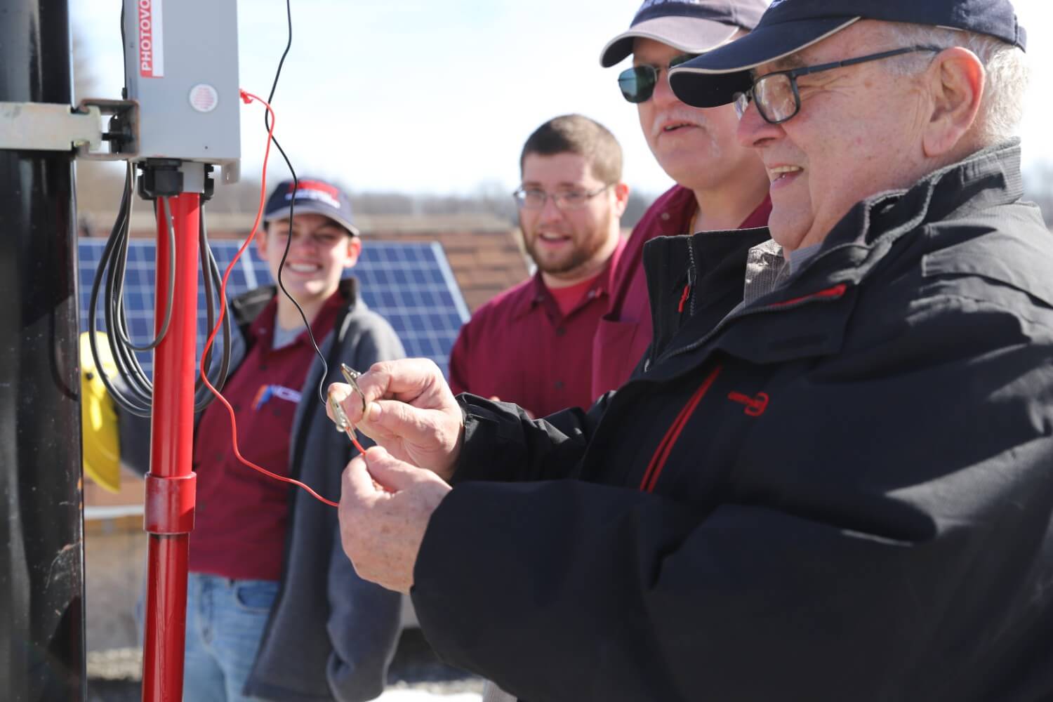 An instructor demonstrating wiring at a photovoltaic panel while NCST students in maroon shirts look on during an outdoor solar training session.